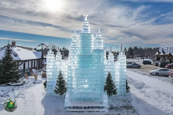 A large, intricate ice sculpture shaped like a castle stands in a snowy parking lot, surrounded by evergreen trees and…