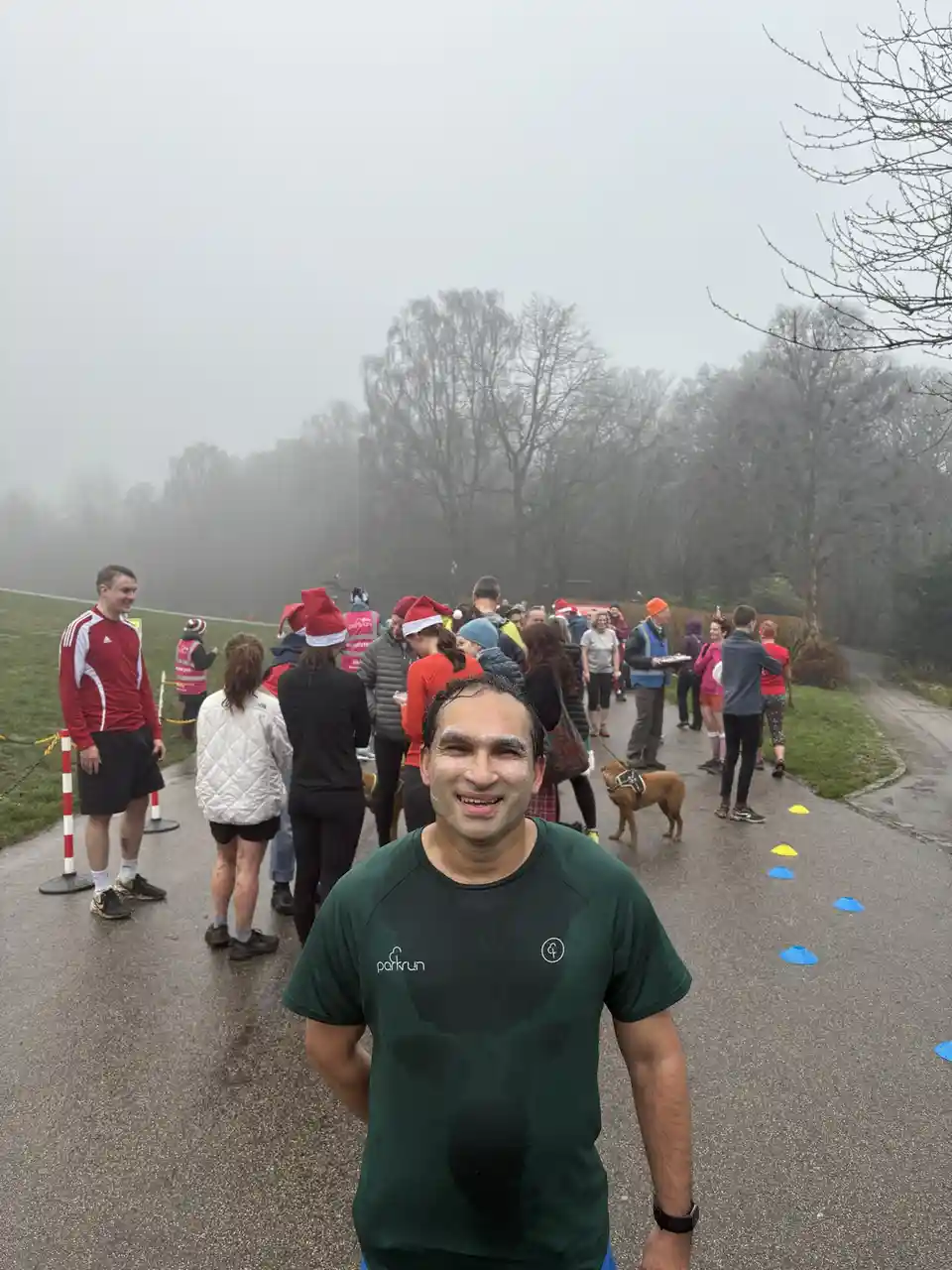 A man in a wet green 'parkrun' t-shirt smiles at the camera after a run, with a foggy park path behind him where other…