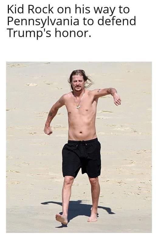 A shirtless man with long hair and tattoos, wearing black swim trunks and a necklace, is walking barefoot on a sandy beach…
