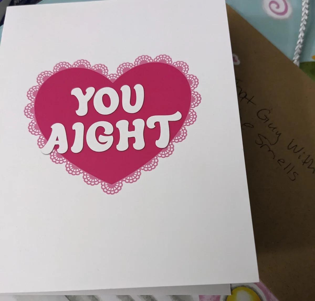 A white greeting card features a large pink heart with a lace-like border, inside which the words 'YOU AIGHT' are written in…
