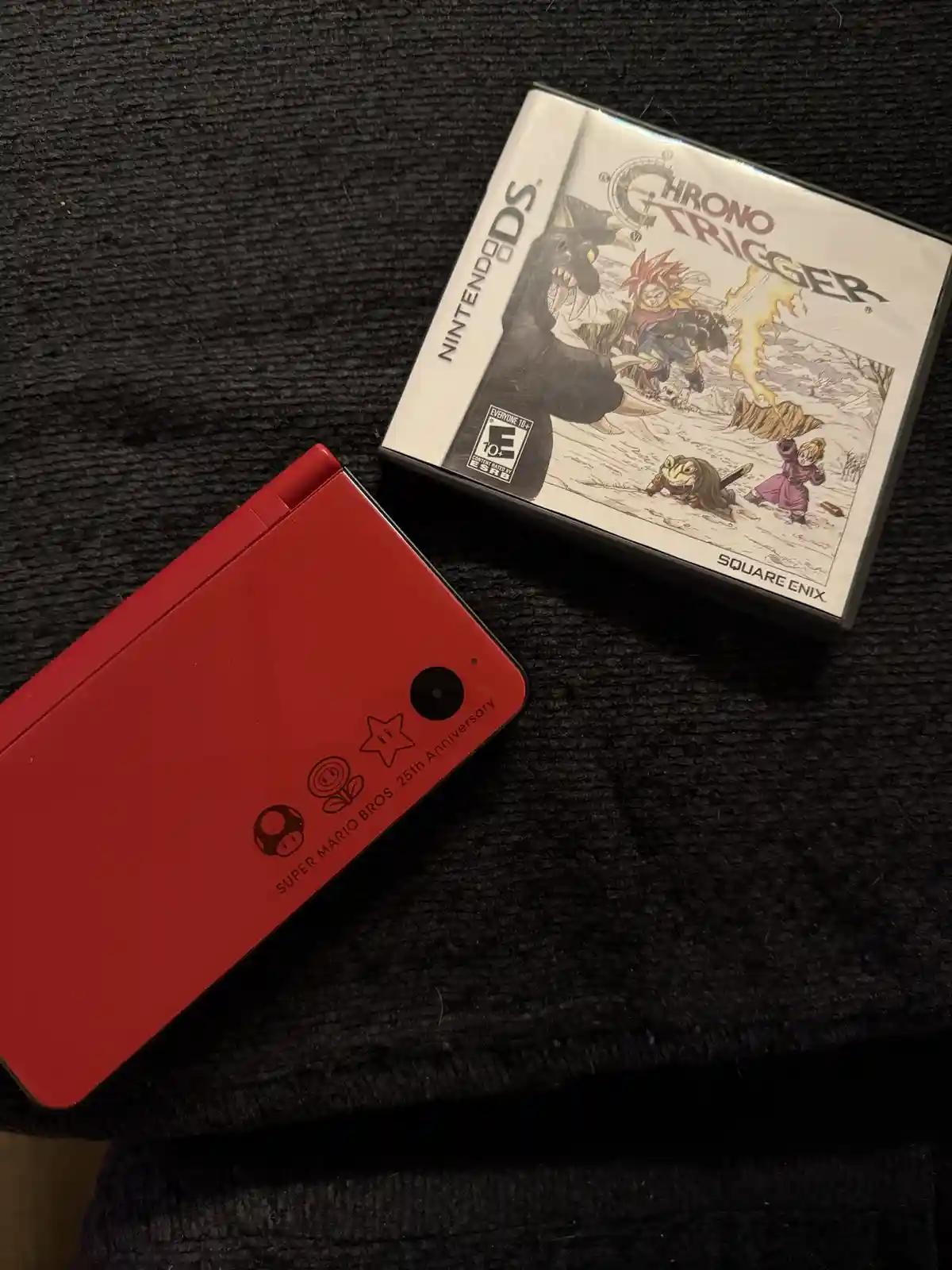 A red Nintendo DS console with 'Super Mario Bros. 25th Anniversary' branding lies next to a sealed Chrono Trigger game box…