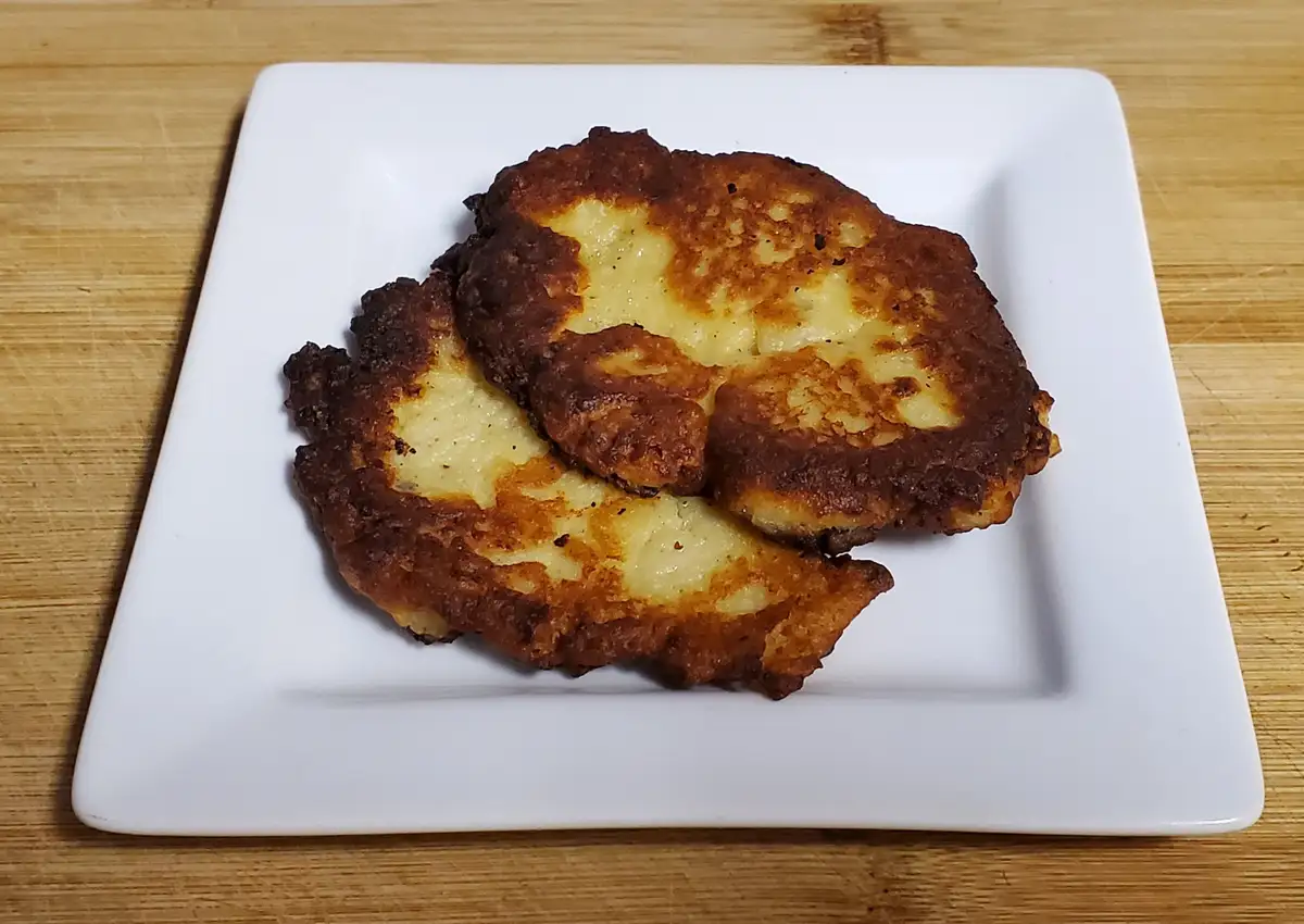 Two golden-brown, slightly burnt pancakes sit on a white square plate atop a wooden surface. The pancakes have a crispy…