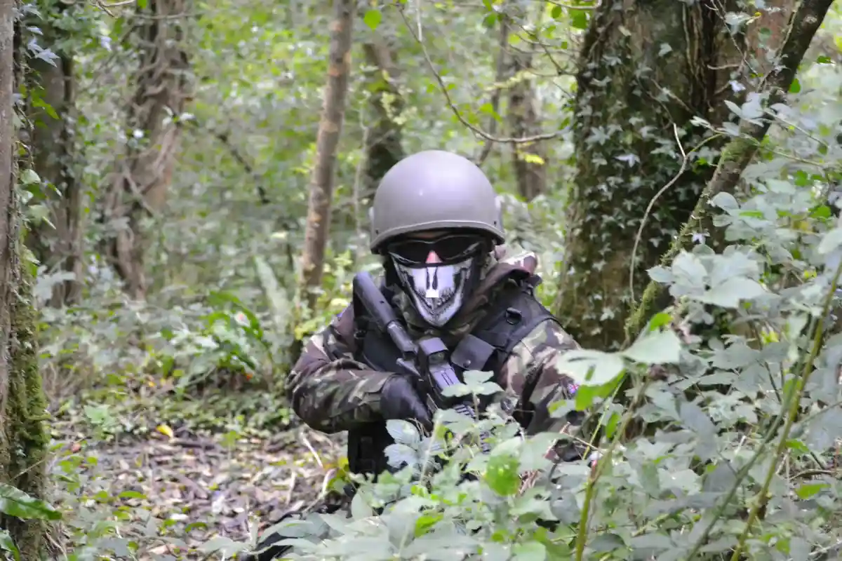 A person in camouflage gear, wearing a grey helmet, black sunglasses, and a white skull-patterned face mask, is crouched in…