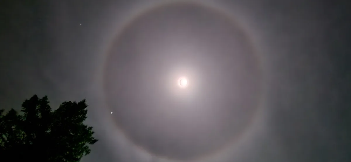 A photograph shows a bright moon centered in a dark, cloudy sky, encircled by a large, faint, circular halo. A dark…