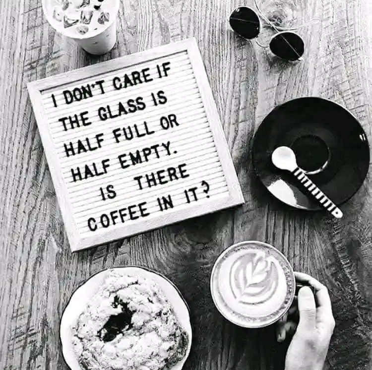 A black-and-white overhead shot of a wooden table with a letterboard sign reading 'I DON'T CARE IF THE GLASS IS HALF FULL OR…