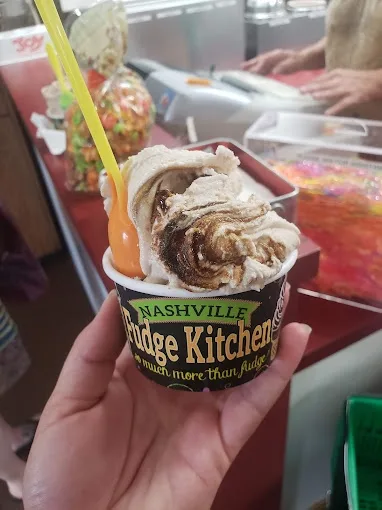 A hand holds a cup of swirled ice cream from 'Nashville Fudge Kitchen' with a yellow spoon and orange plastic spoon inside…