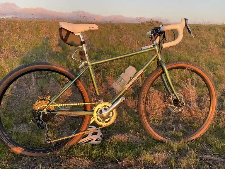 A green bicycle with white handlebars and a white saddle is parked in a grassy field during sunset. A water bottle is…