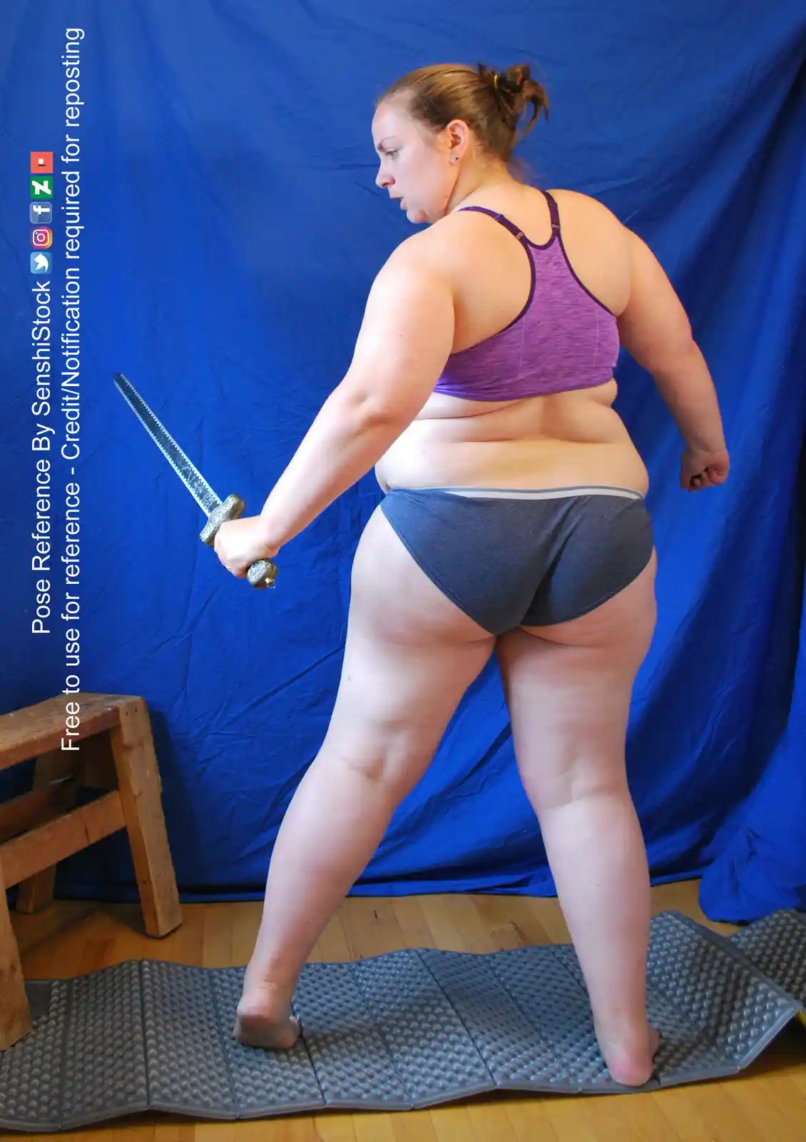 A woman in a purple sports bra and grey underwear stands in a warrior pose, holding a sword, against a blue backdrop. She is…
