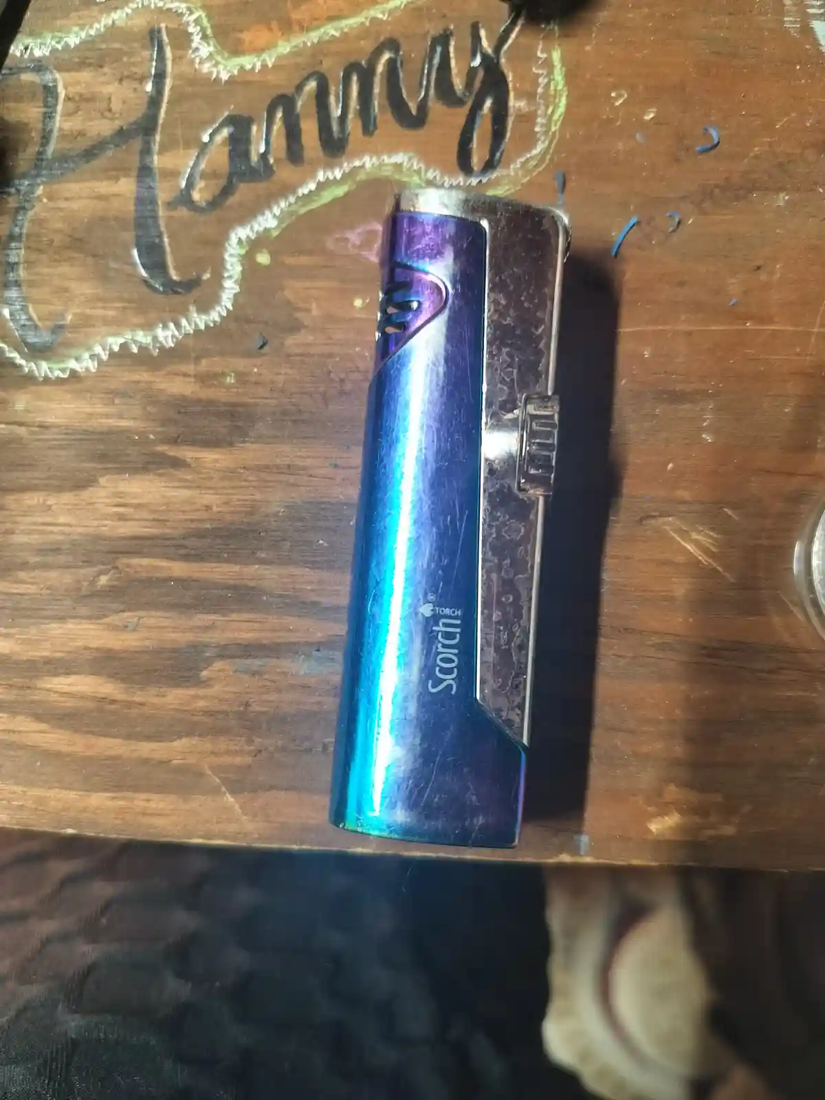 A blue and purple iridescent Scorch Torch lighter rests on a worn wooden table with the name 'Hannys' written in chalk…