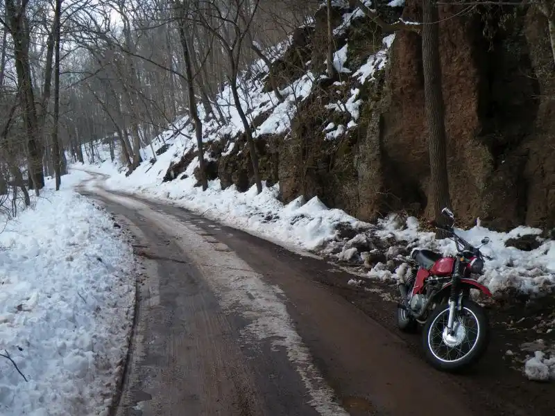 A red dirt bike is parked on the side of a snowy, winding forest road with snow-covered banks and bare trees. The overlaid…
