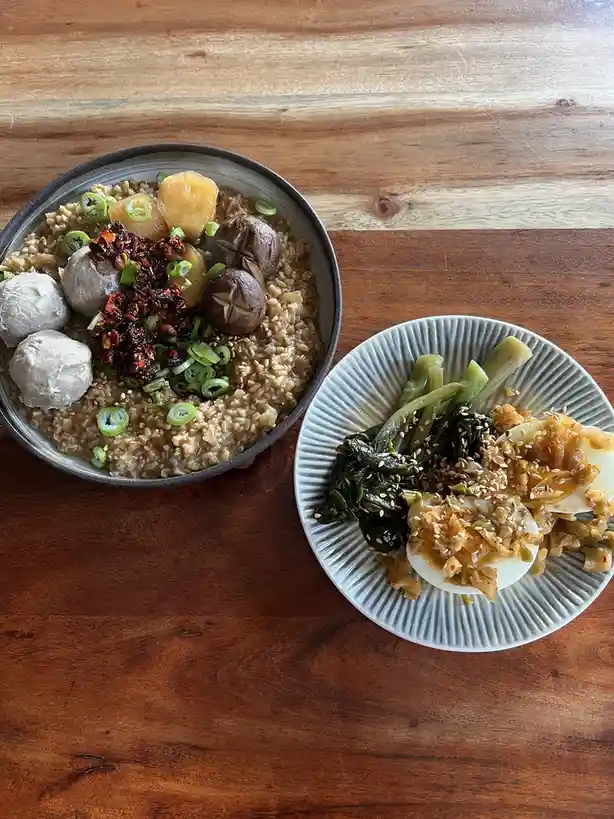 A top-down photo shows a bowl of brown rice topped with meatballs, mushrooms, potato chunks, green onions, and red chili…