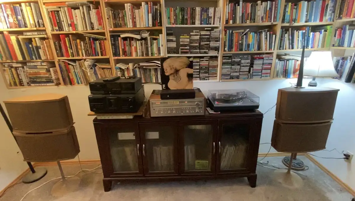 A room with wooden bookshelves filled with books and CDs, a dark wooden media console holding a record player, stereo…