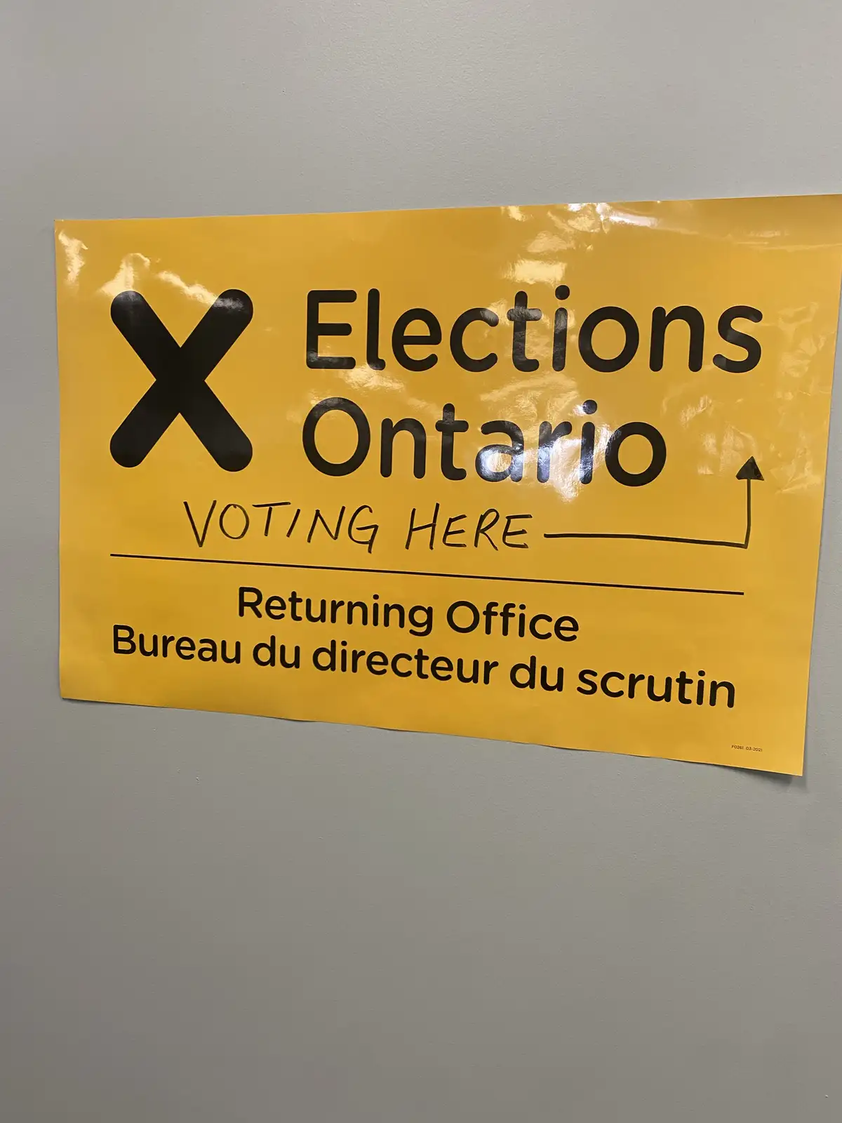A yellow Elections Ontario sign taped to a gray wall indicates a voting location with printed text and a handwritten arrow…