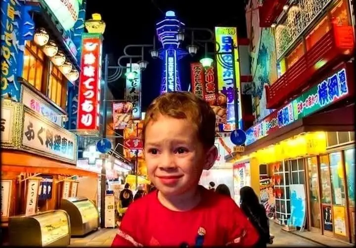 A young boy with a pouty, tearful expression is superimposed over a brightly lit, colorful Japanese street at night, filled…
