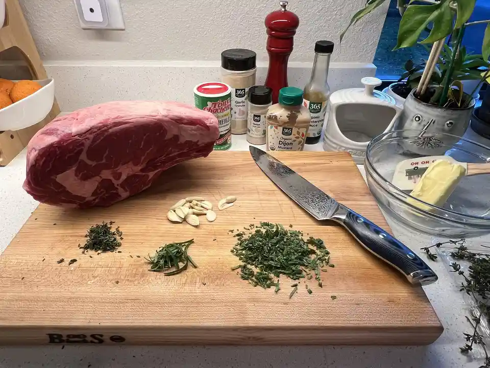 A large raw beef roast sits on a wooden cutting board next to chopped herbs, garlic, and a knife, with various seasonings…