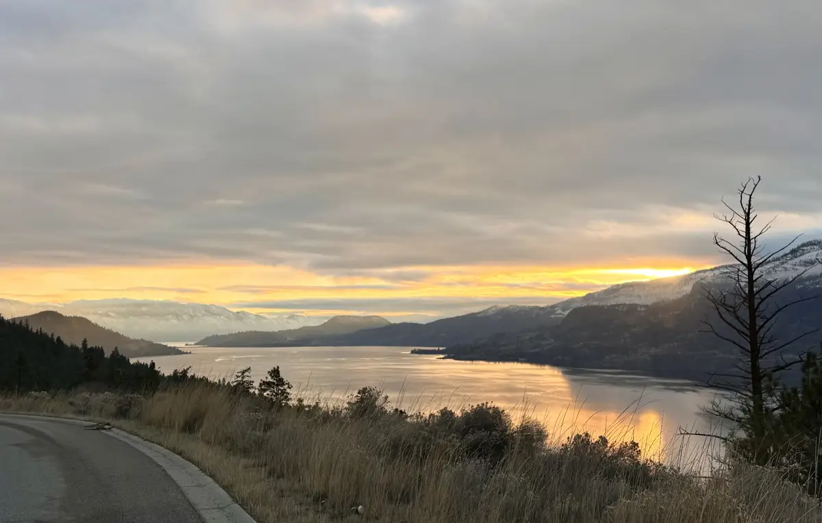 A scenic photograph captures a serene sunset over a large lake surrounded by snow-capped mountains, with a winding road in…
