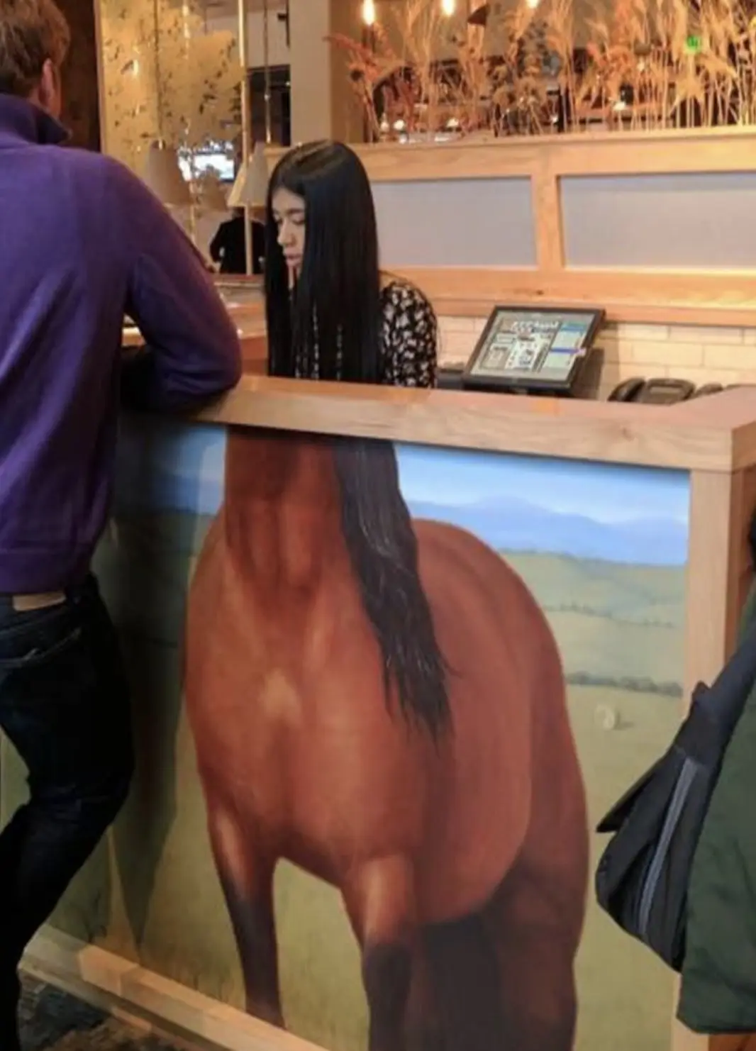 A person with long black hair is standing behind a counter, looking down, while a large painting of a horse's rear end is…