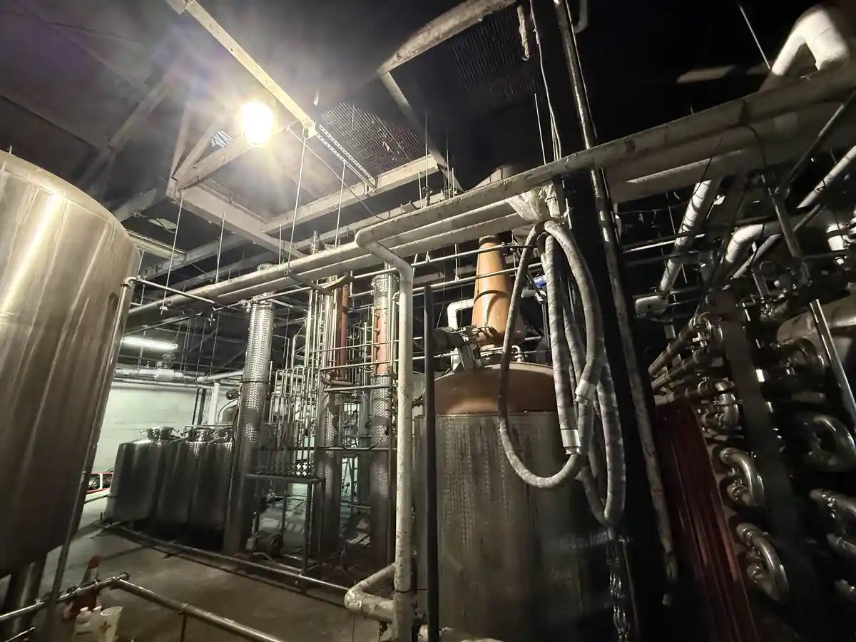 A dimly lit industrial distillery interior with large stainless steel tanks, copper stills, and an intricate network of…