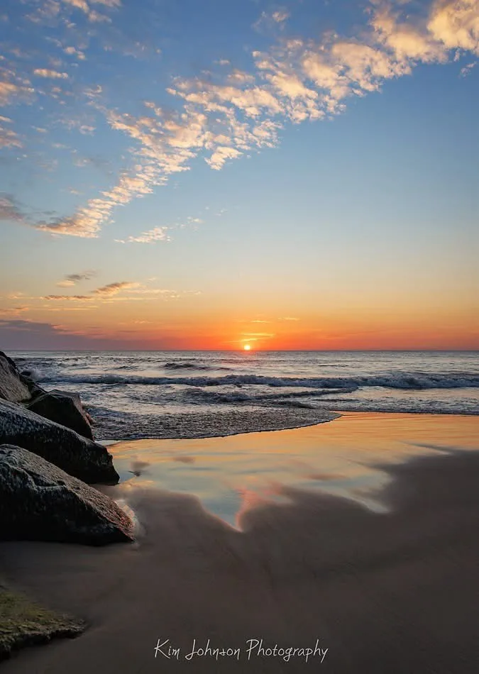 A serene beach sunset photo with orange and blue skies, waves lapping at wet sand, and large rocks in the foreground. The…