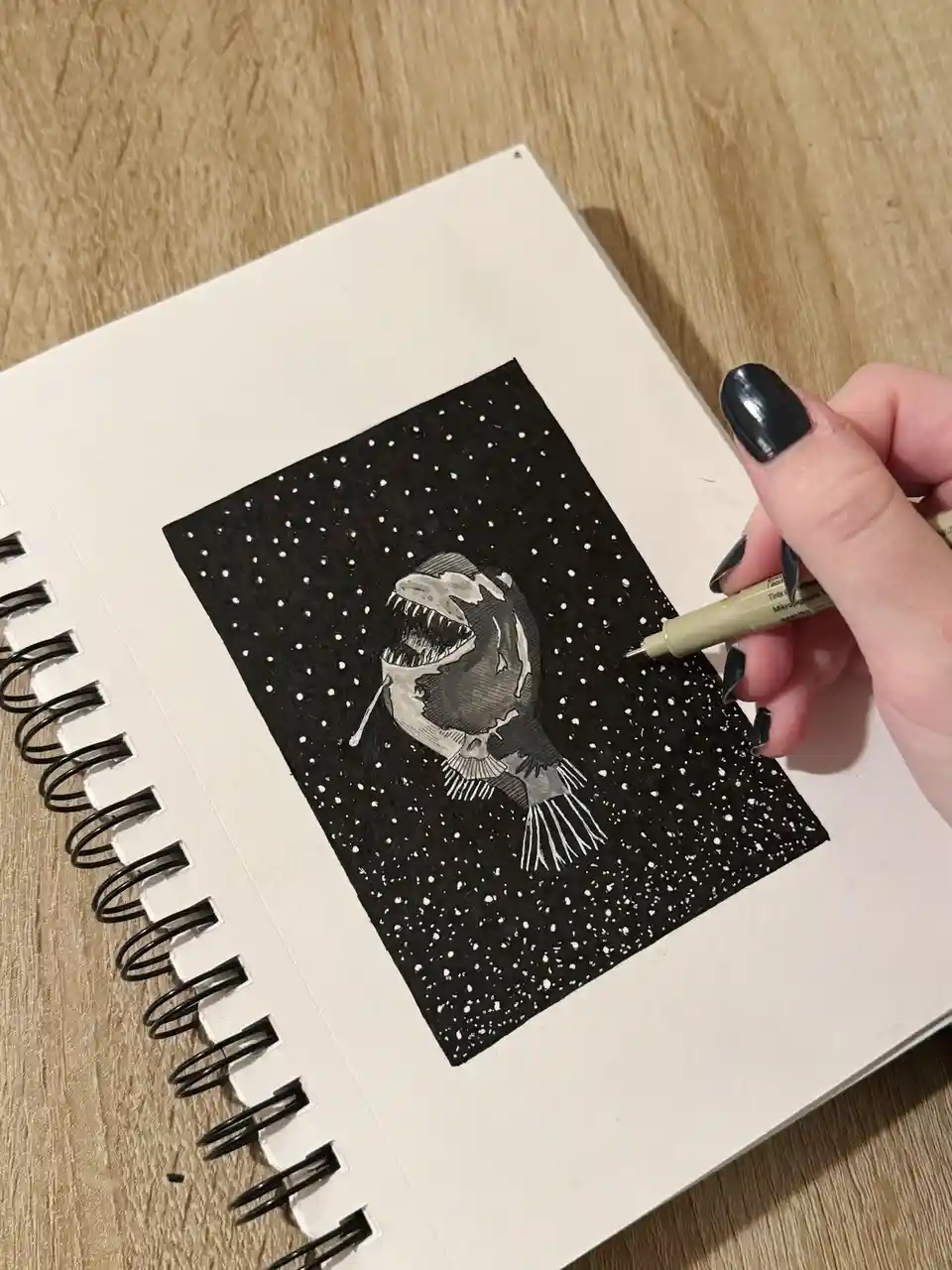A hand with black nail polish holds a light green pen over a sketchbook page, which features a detailed black-and-white…