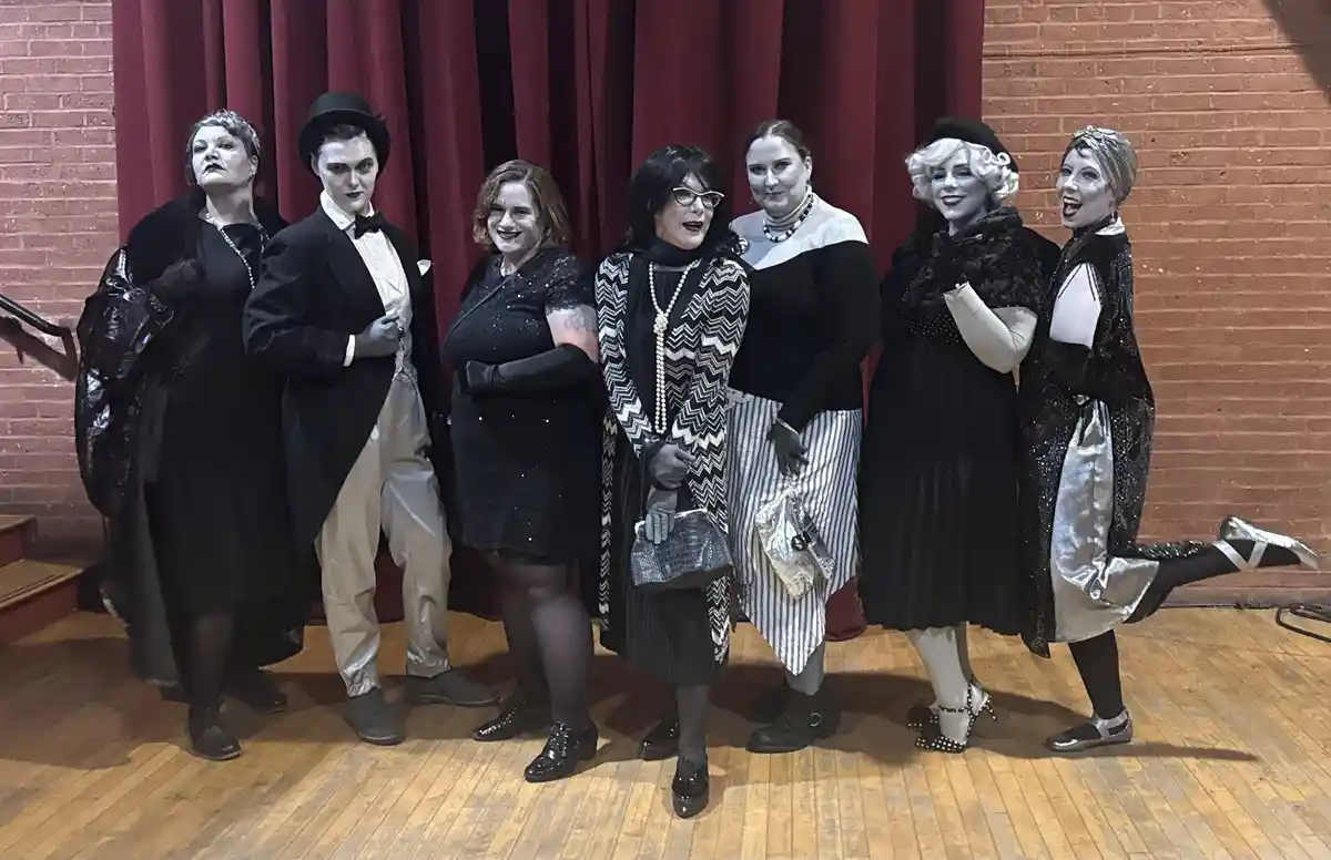 Seven people in theatrical makeup and costumes pose on a wooden stage in front of red curtains and brick walls. Their faces…
