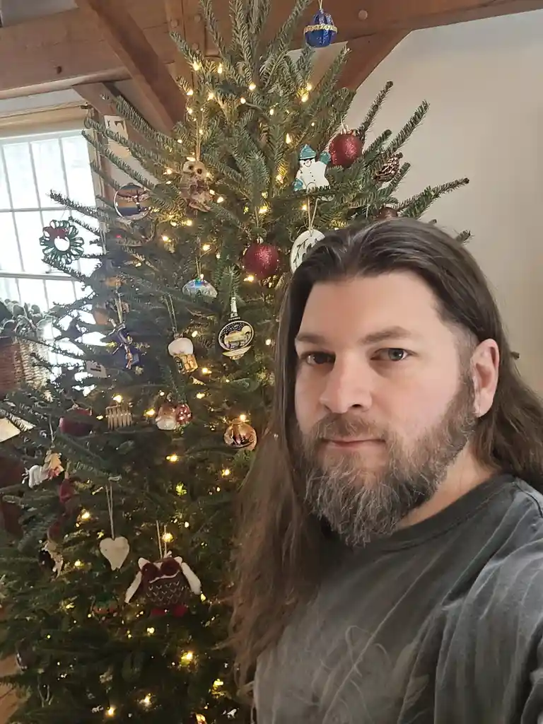 A man with long brown hair and a beard stands in front of a decorated Christmas tree with lights and ornaments. The tree is…