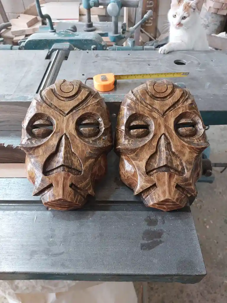 Two intricately carved wooden masks with stylized eyes and noses sit side by side on a metal workbench in a workshop. Behind…