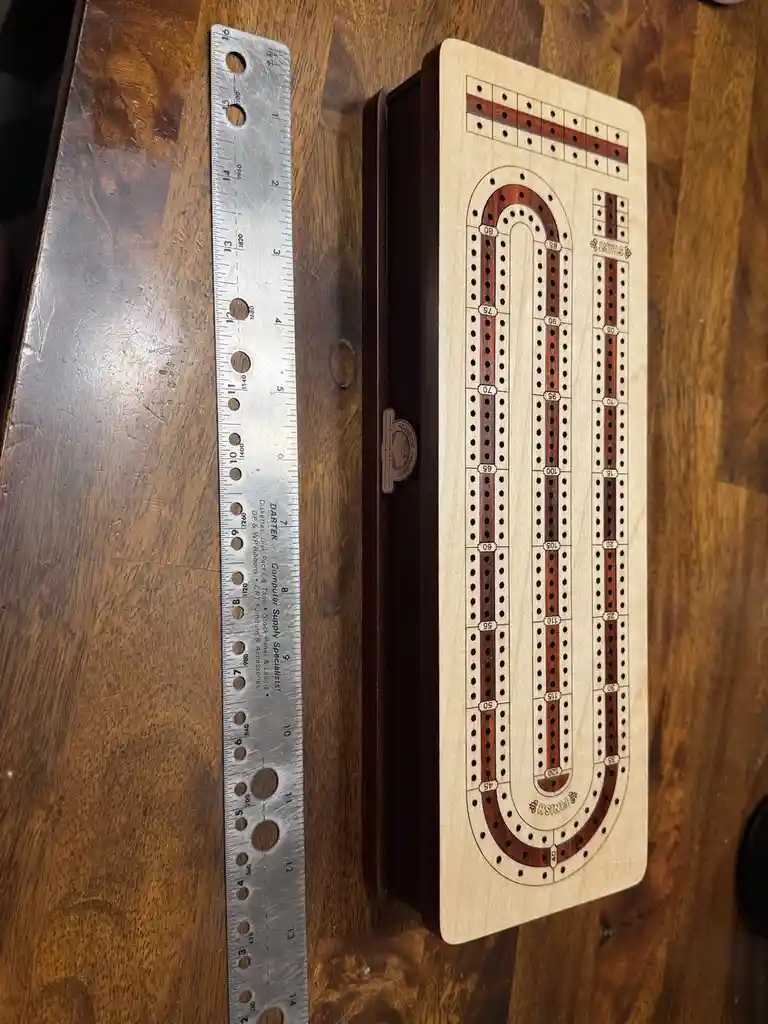 A wooden cribbage board with a U-shaped track and numbered scoring holes lies next to a metal ruler on a wooden table…