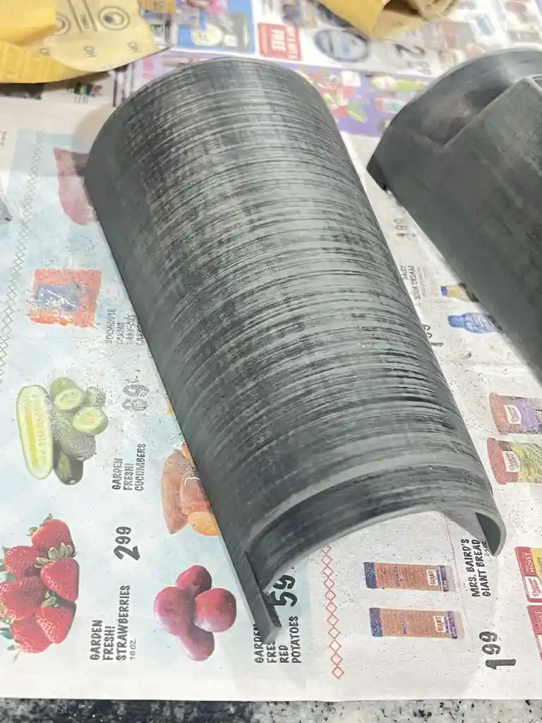 A close-up photo shows two black-painted, curved plastic or metal parts resting on a grocery store flyer featuring images of…