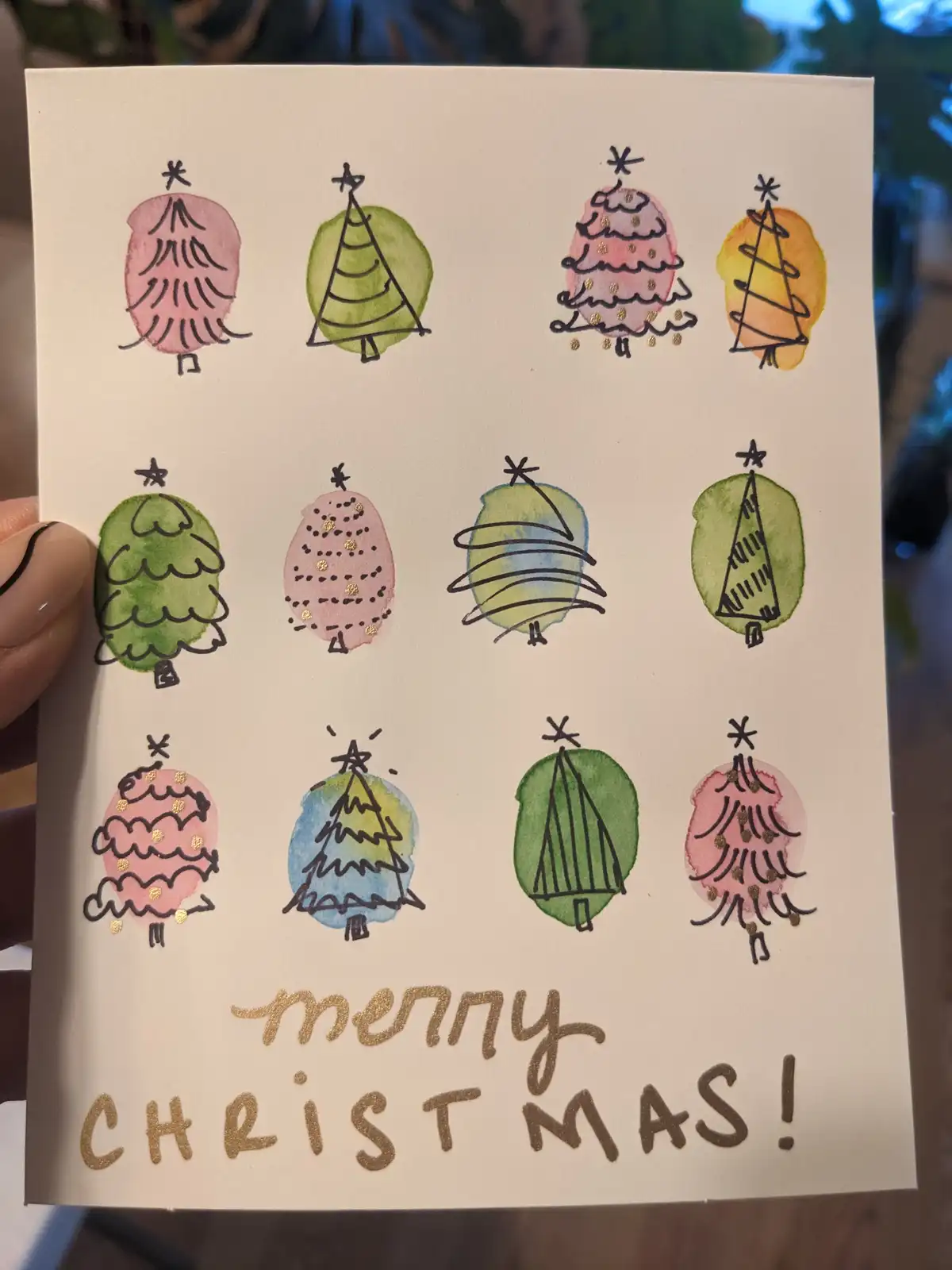 A hand holds a handmade Christmas card featuring twelve distinct, whimsically drawn Christmas trees in various shapes…