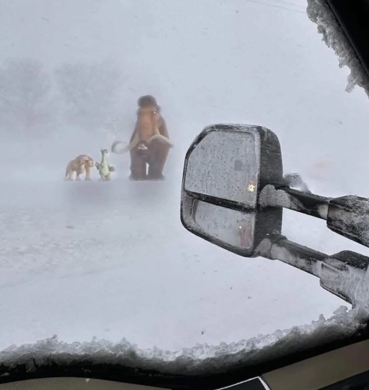 A snowy, frost-covered car windshield view shows three small, toy-like figures resembling characters from Ice Age: a…