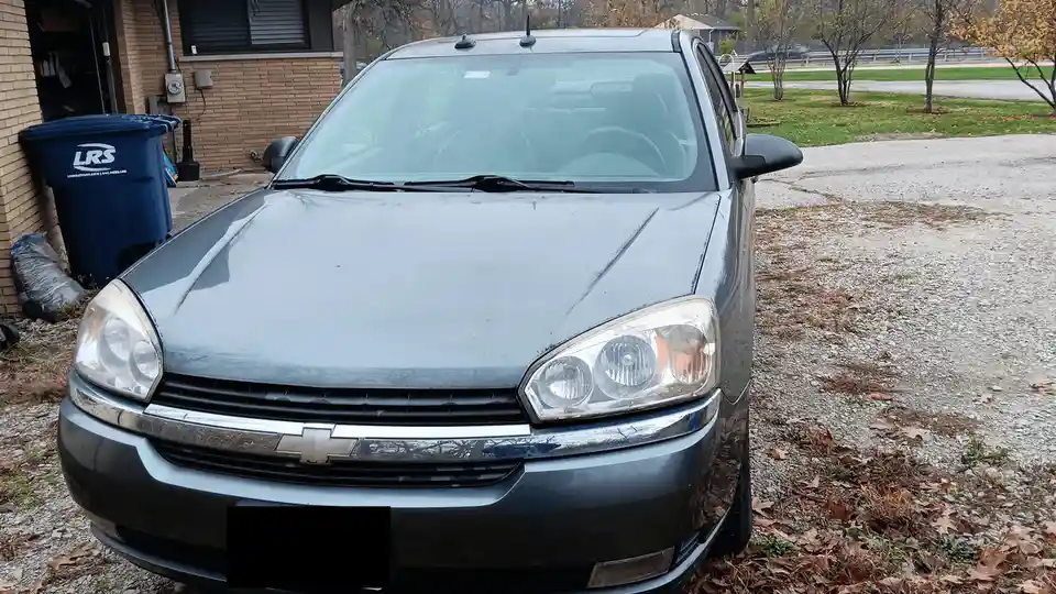 A gray Chevrolet sedan is parked on a gravel driveway in front of a brick house, with autumn leaves scattered around. The…