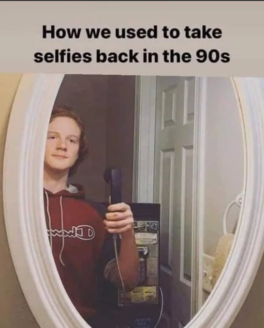 A young person with reddish hair wearing a red Champion hoodie holds a black corded telephone receiver up to a mirror…