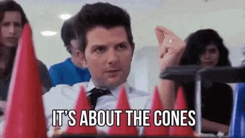 A man in a white shirt and tie sits at a table with red traffic cones in front of him, looking confused or exasperated…