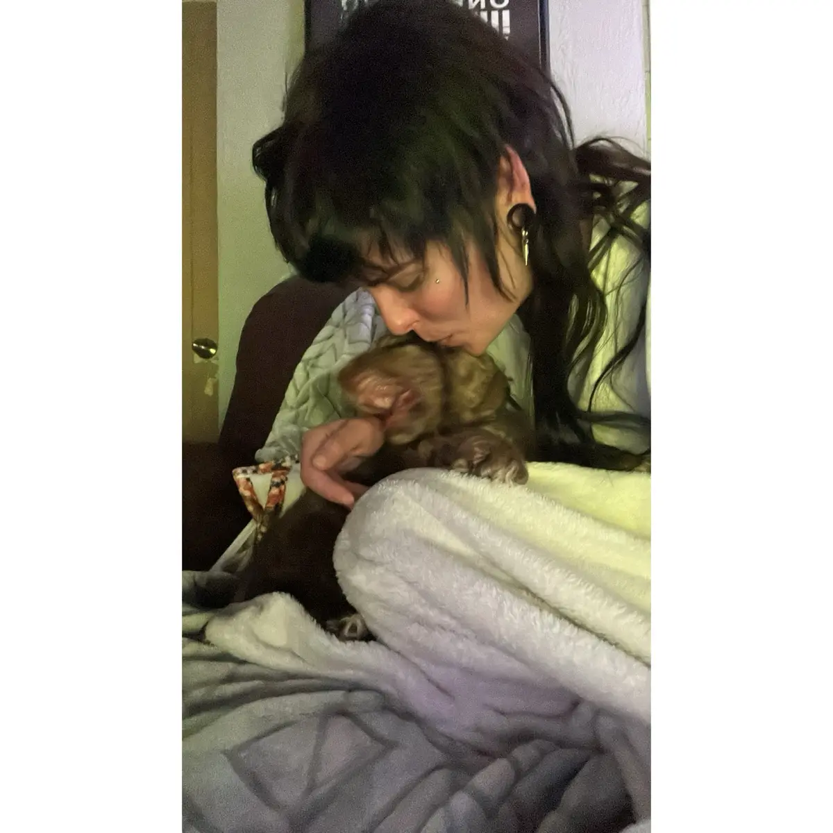 A person with dark hair and visible ear piercings is shown leaning down to kiss a small, fluffy brown puppy that is nestled…