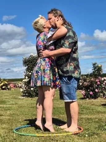 A man and woman are kissing passionately in a sunny garden with colorful flowers and a blue sky. A rainbow-colored hula hoop…