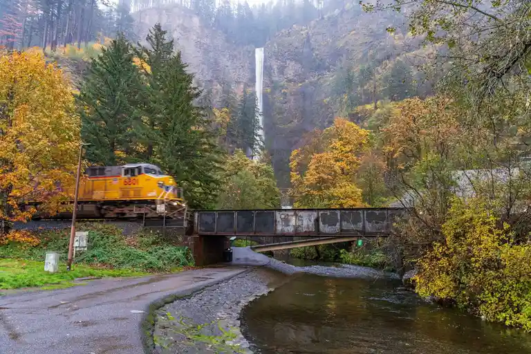 A yellow train with number 6801 on its side is crossing a metal bridge over a river, with a large waterfall visible in the…
