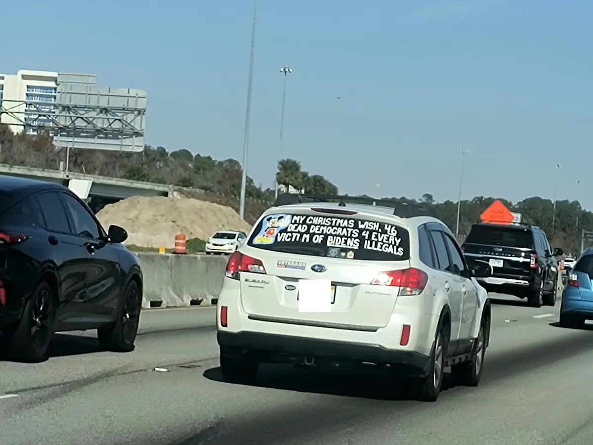 A white Subaru Outback is seen from behind on a highway, its rear window covered in white text that reads 'MY CHRISTMAS…