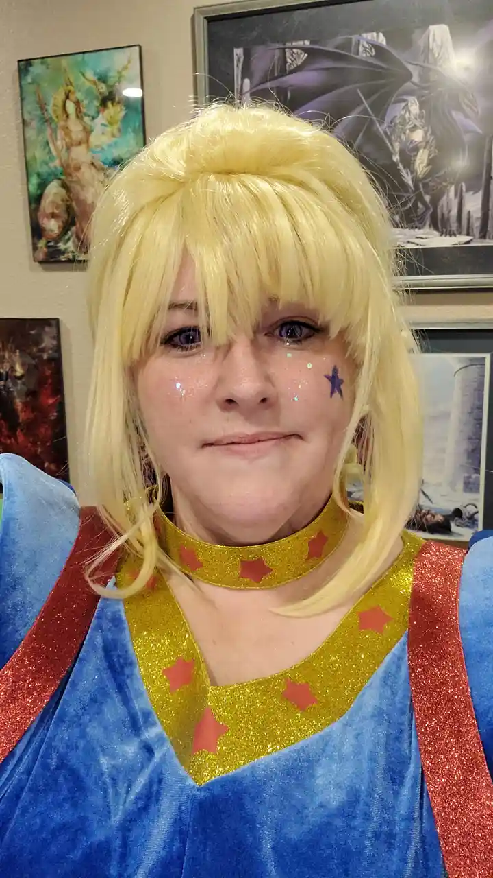 A person in a cosplay costume with a blonde wig, purple eyes, glitter on their cheeks, and a blue, gold, and red outfit with…