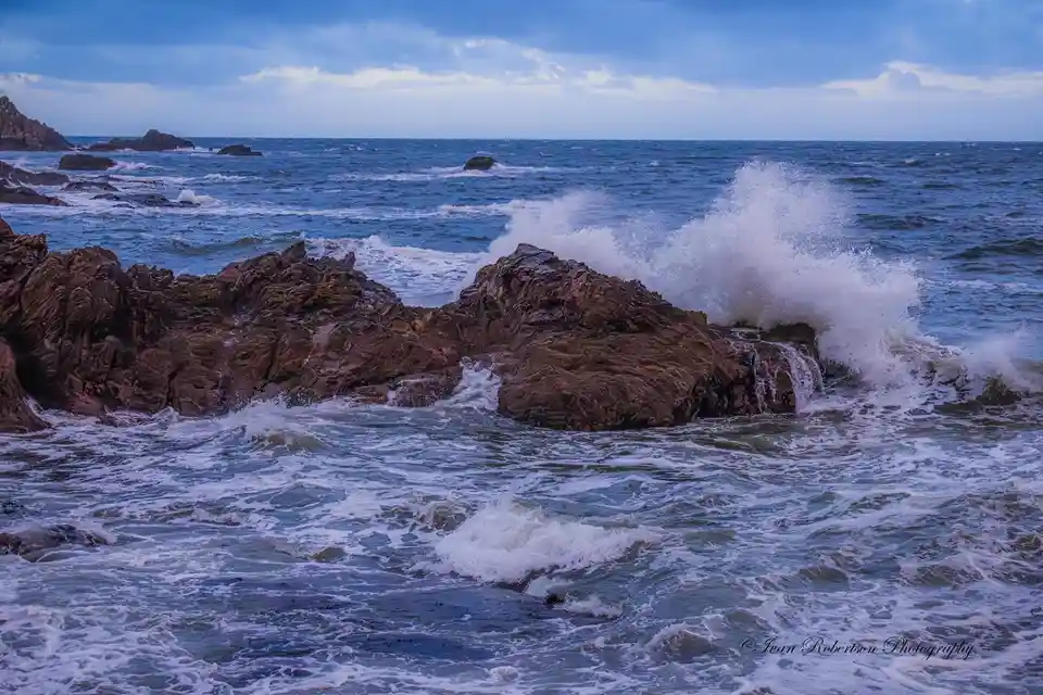 A dramatic photograph shows powerful ocean waves crashing against rugged, reddish-brown coastal rocks, sending white spray…