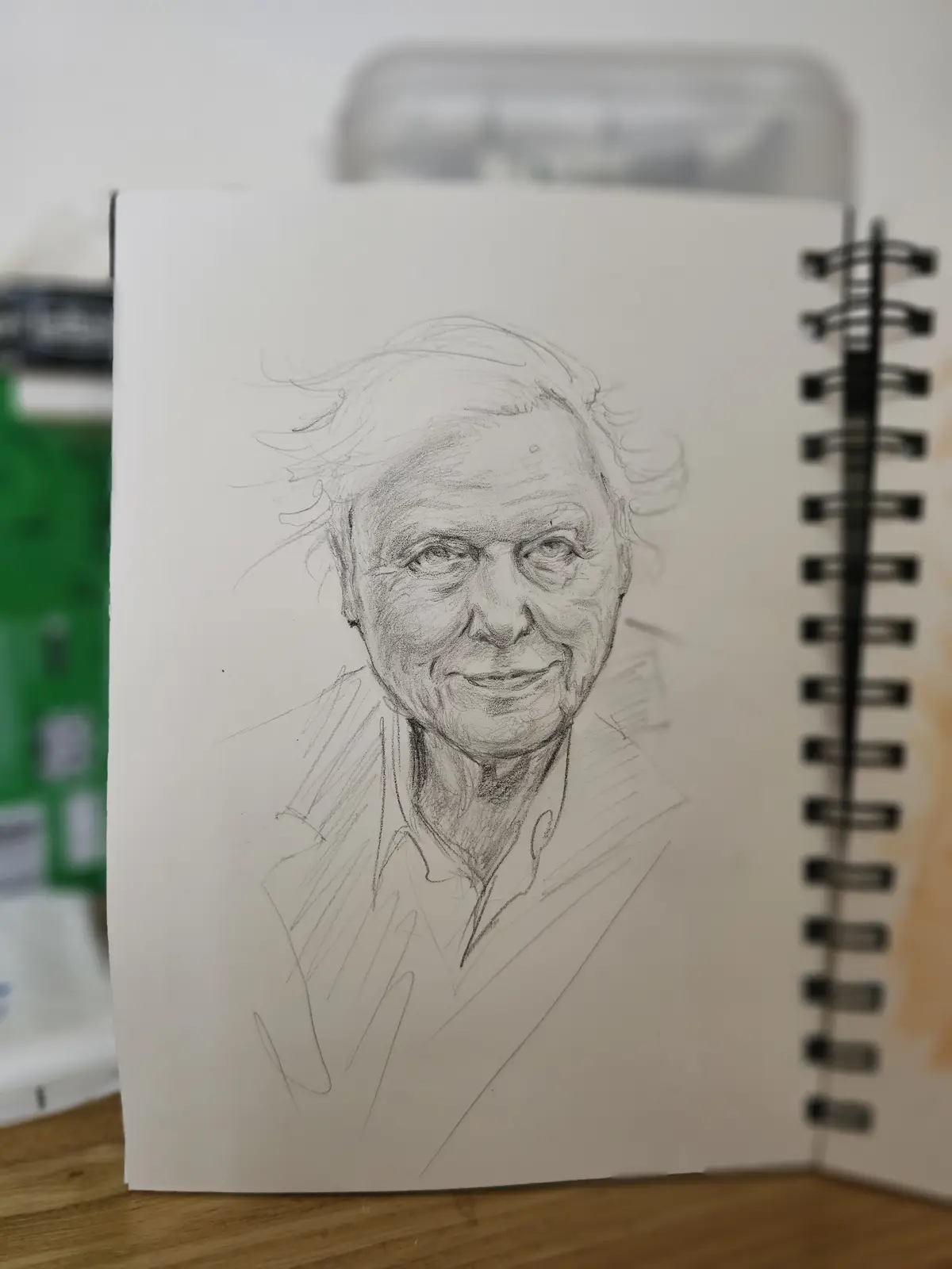 A pencil sketch of an elderly man with wild hair and a warm smile is drawn in a spiral-bound sketchbook. The drawing is…