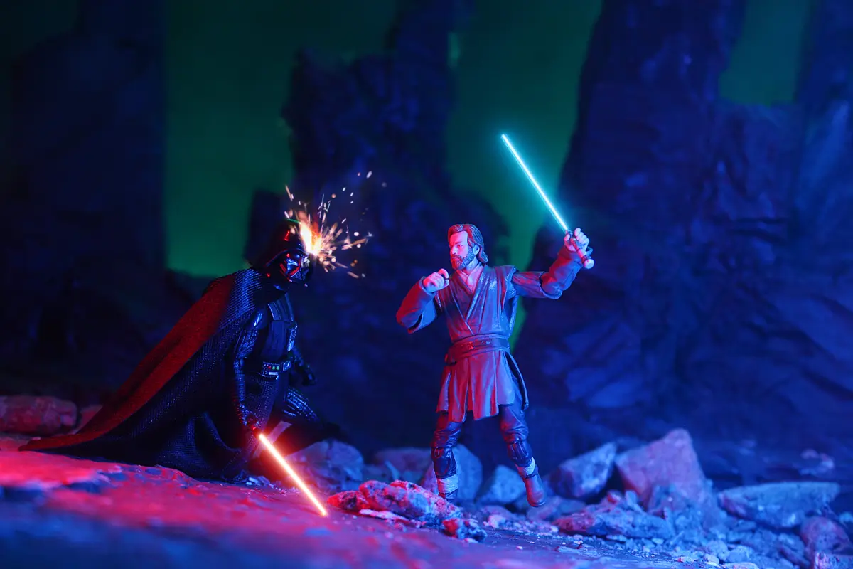 Two Star Wars action figures, Darth Vader with a red lightsaber and Obi-Wan Kenobi with a blue lightsaber, are locked in…