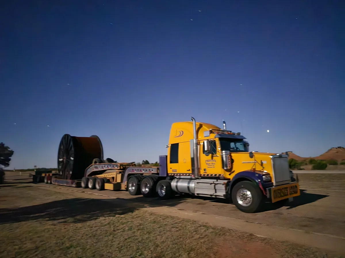 A bright yellow semi-truck with a purple bumper is hauling an enormous, dark-colored spool on a flatbed trailer under a…