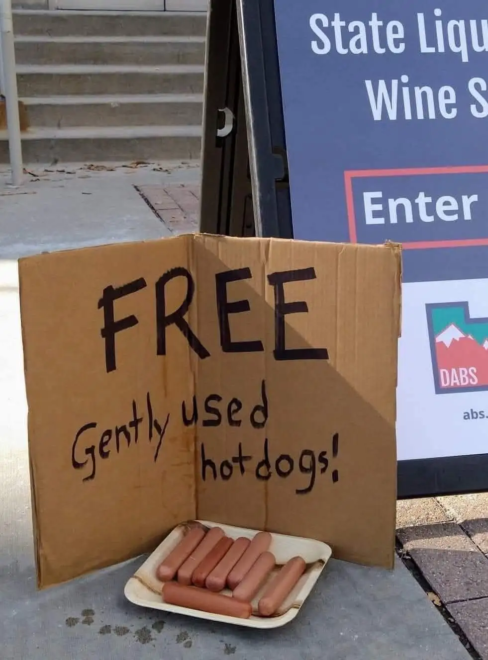 A handmade cardboard sign on a sidewalk reads 'FREE Gently used hotdogs!' and sits beside a tray of uncooked hot dogs…