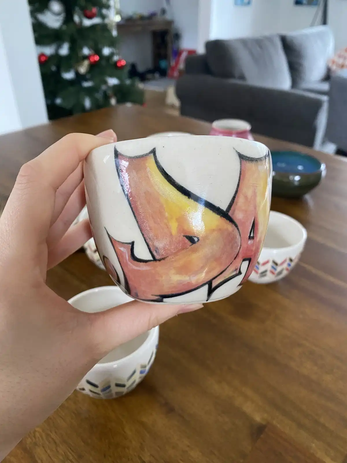 A hand holds up a handmade ceramic mug painted with a stylized orange and yellow fox face, outlined in black. The mug is…
