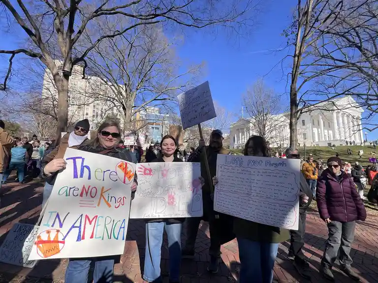 A group of protesters stands outdoors in front of a large white building under a clear blue sky, holding handmade signs. One…