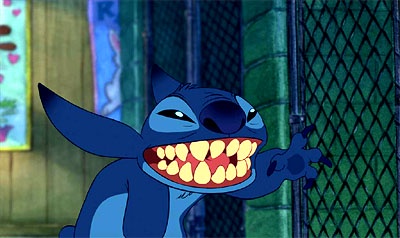 A blue cartoon alien with large ears and sharp teeth is grinning widely while leaning against a chain-link fence. The…