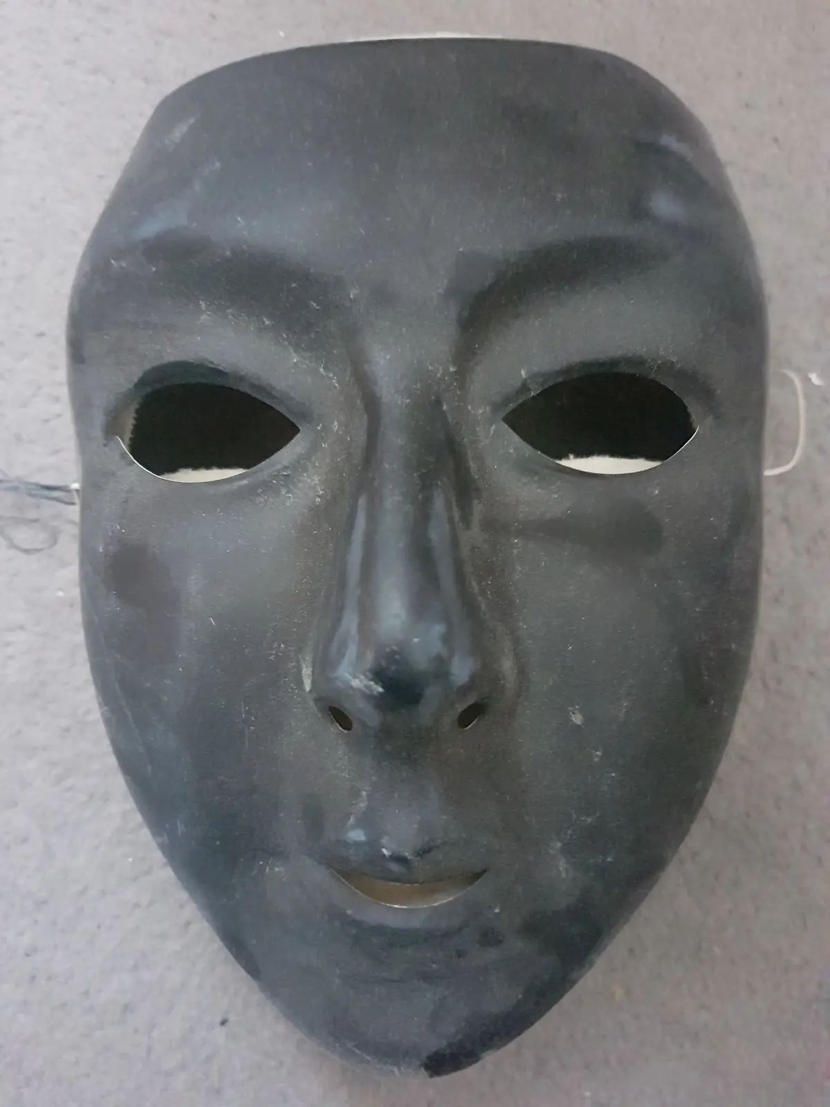 A plain black, featureless mask with eye, nose, and mouth holes lies on a textured gray surface. The overlaid text reads…