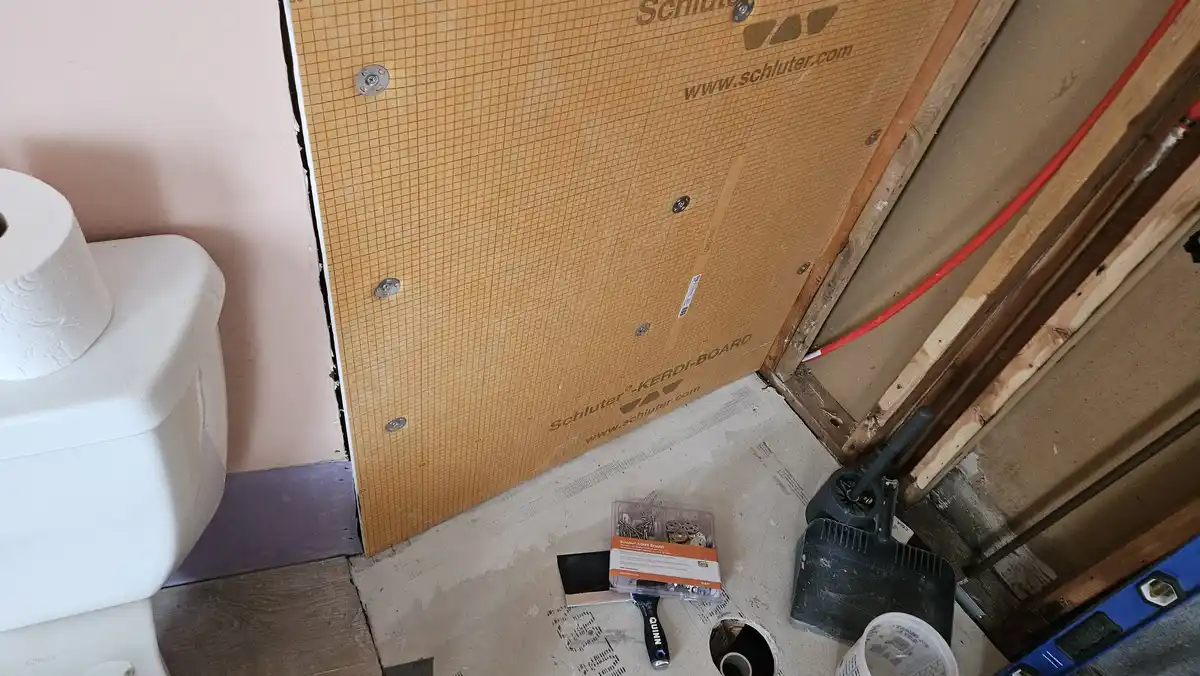 A bathroom under renovation shows a toilet on the left next to a wall with Schluter-KERDI-BOARD installed, tools and…