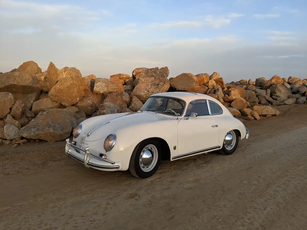 A pristine white vintage Porsche 356 coupe is parked on sandy ground in front of a large pile of rough, brownish-orange…
